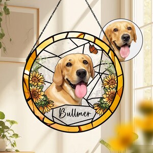 Op de afbeelding: Een rond gebrandschilderd raam met een gouden Labrador Retriever met zijn tong uit, omringd door zonnebloemen en een vlinder. De naam "Bullmer" staat eronder. Het raam heeft een gele rand en hangt aan een zwarte ketting.