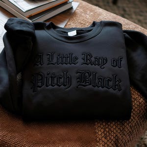 May include: Black sweatshirt with the phrase "A Little Ray of Pitch Black" embroidered in gothic lettering. The sweatshirt is folded and resting on a patterned surface, with a stack of books visible in the background.