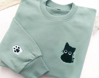 Op de afbeelding: Een lichtgroen sweatshirt met een zwarte kat en een witte voetafdruk geborduurd erop.