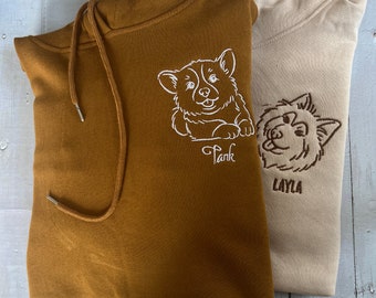 May include: Two brown hooded sweatshirts with white embroidered dog portraits. The sweatshirt on the left has a corgi with the name "Tank" embroidered below it. The sweatshirt on the right has a fluffy dog with the name "Layla" embroidered below it.