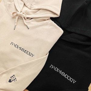 Puede incluir: Dos sudaderas con capucha, una beige y otra negra, con la inscripción en números romanos "IV.IX.MMXXXIV" en cada una. La sudadera beige tiene un pequeño corazón con las letras "EF" bordadas en la manga.