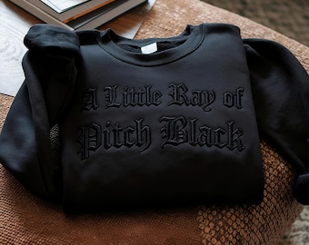 May include: A black sweatshirt with the phrase "A Little Ray of Pitch Black" embroidered in a gothic font. The sweatshirt is folded on a brown surface, with a neutral background.