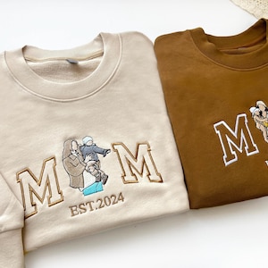 May include: Two sweatshirts with embroidered "MOM" lettering and family photos. The beige sweatshirt has the text "EST. 2024" and "Grace Olivia" on the sleeve. The brown sweatshirt has the text "EST. 2024" and a family photo of three people.