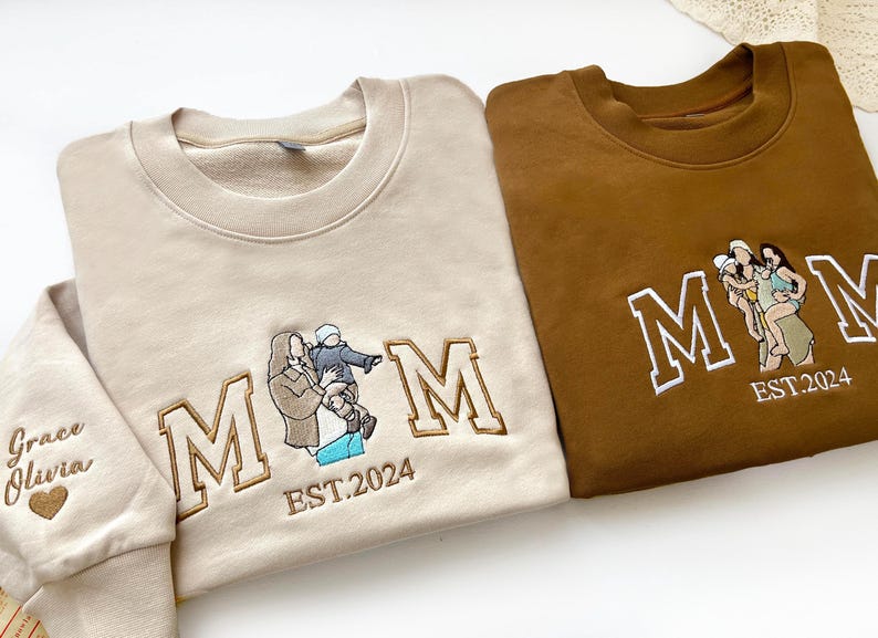 May include: Two sweatshirts with embroidered "MOM" lettering and family photos. The beige sweatshirt has the text "EST. 2024" and "Grace Olivia" on the sleeve. The brown sweatshirt has the text "EST. 2024" and a family photo of three people.