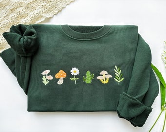 May include: A dark green sweatshirt with embroidered images of a mushroom, flower, cactus, and mushrooms. The embroidery is in shades of orange, yellow, white, and green.