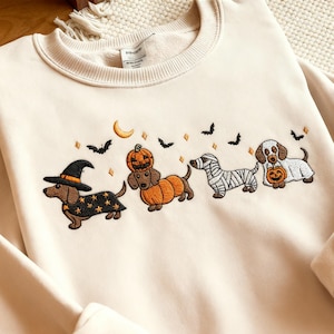 May include: Cream-colored sweatshirt with an embroidered Halloween dachshund design. The design features four dachshunds dressed in costumes: a witch, a pumpkin, a mummy, and a dog holding a pumpkin. Bats, a crescent moon, and diamonds complete the festive look.