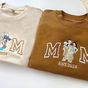 May include: Two sweatshirts with embroidered "MOM" text and a family image. The sweatshirt on the left is beige with a family of two, and the sweatshirt on the right is brown with a family of three.
