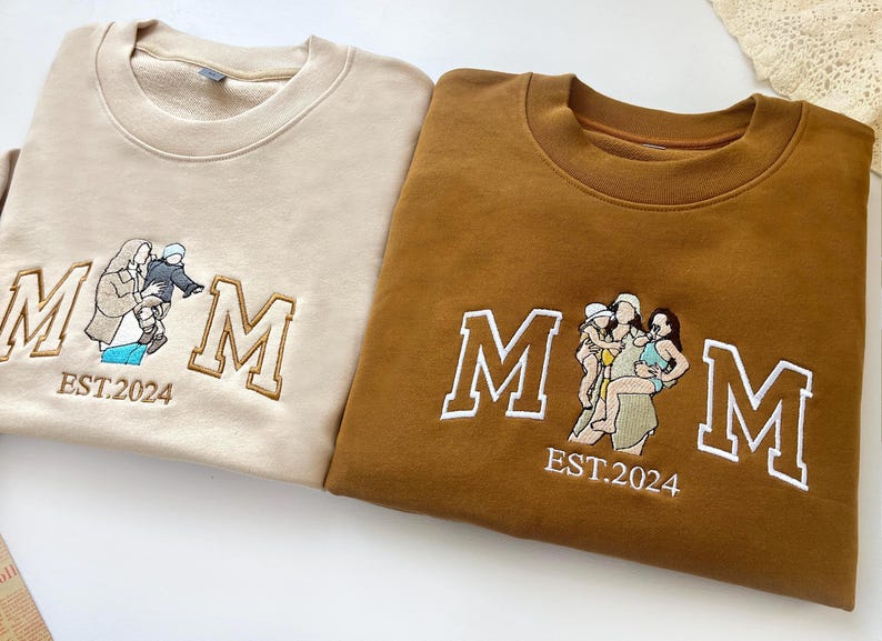 May include: Two sweatshirts with embroidered "MOM" text and a family image. The sweatshirt on the left is beige with a family of two, and the sweatshirt on the right is brown with a family of three.