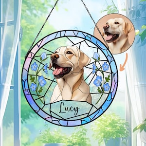 Op de afbeelding: Een ronde glas-in-lood raam met een Labrador Retriever, blauwe bloemen en de naam "Lucy". Het raam is omlijnd met zwarte lijnen en hangt aan een ketting. Een kleinere afbeelding van de hond staat rechtsboven.