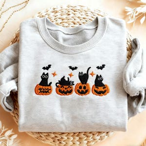 Peut inclure: Un sweat-shirt gris clair avec une broderie sur le thème d'Halloween. Le motif comprend quatre chats noirs assis dans des citrouilles orange, avec des chauves-souris et des étoiles au-dessus. Le sweat-shirt est en matière douce.