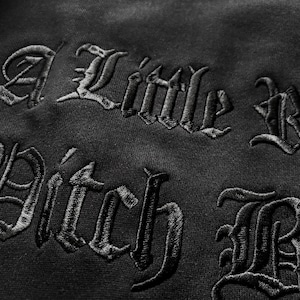 May include: Close-up of black fabric with embroidered text in a gothic font. The text reads "A Little Bad Pitch Black". The embroidery is detailed and the fabric has a subtle texture.