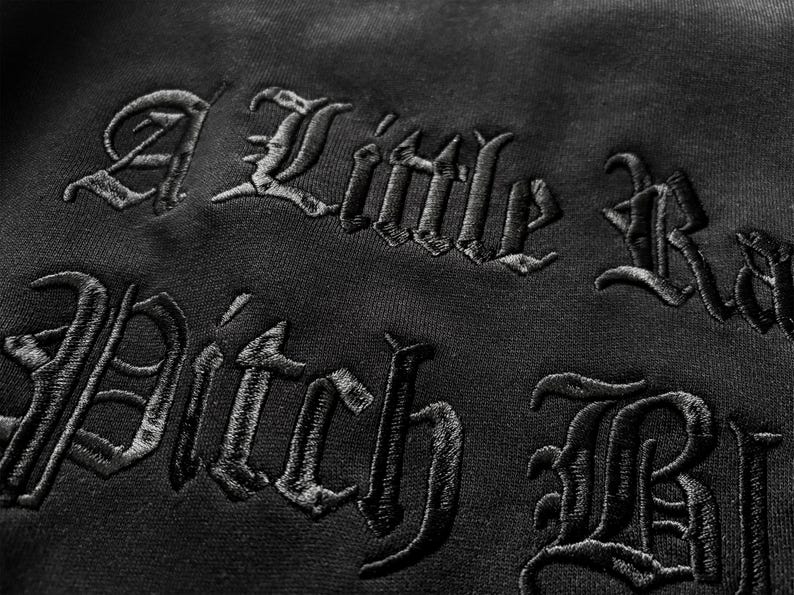 May include: Close-up of black fabric with embroidered text in a gothic font. The text reads "A Little Bad Pitch Black". The embroidery is detailed and the fabric has a subtle texture.