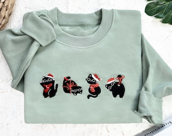 May include: A pale green sweatshirt with an embroidered design of four black cats in Christmas outfits. Each cat has a different festive accessory, such as Santa hats and scarves. The sweatshirt has a round neck and long sleeves.