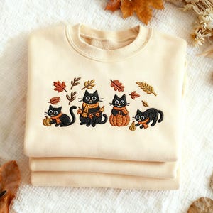 Peut inclure: Un sweat-shirt crème avec une broderie de quatre chats noirs avec des écharpes oranges et des citrouilles, entourés de feuilles d'automne. Le sweat-shirt est plié, mettant en valeur le motif sur la couche supérieure.