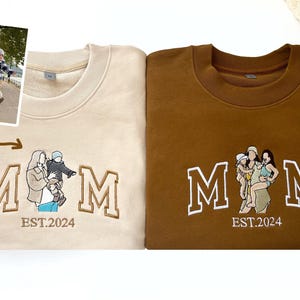 May include: Two beige and brown sweatshirts with embroidered white letters spelling "MOM" and a small embroidered image of a woman and two children. The text "EST. 2024" is embroidered below the letters.