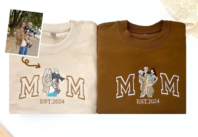 May include: Two beige and brown sweatshirts with embroidered white letters spelling "MOM" and a small embroidered image of a woman and two children. The text "EST. 2024" is embroidered below the letters.
