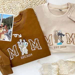 May include: Two brown and beige sweatshirts with embroidered "MOM" lettering. The sweatshirts feature a family portrait with the text "EST. 2024" underneath. One sweatshirt has the names "Grace Olivia" embroidered on the sleeve.