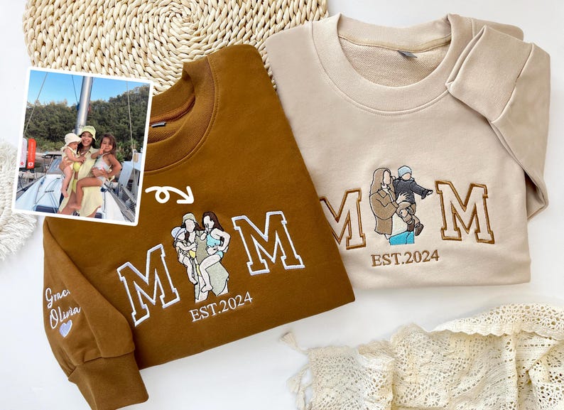 May include: Two brown and beige sweatshirts with embroidered "MOM" lettering. The sweatshirts feature a family portrait with the text "EST. 2024" underneath. One sweatshirt has the names "Grace Olivia" embroidered on the sleeve.
