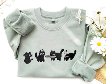 May include: A light green sweatshirt with black embroidered cats. The cats are in a line and one is holding a knife. The sweatshirt has a crew neck and ribbed cuffs.