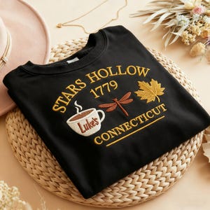 May include: Black crew neck sweatshirt with embroidered text and graphics. The design includes "STARS HOLLOW 1779 CONNECTICUT", a coffee cup with "Luke's", a dragonfly, and a maple leaf. The sweatshirt is displayed on a woven mat.