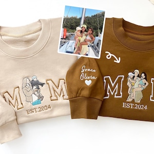 May include: Two beige and brown sweatshirts with the word "MOM" embroidered in a large font. The beige sweatshirt has the text "Grace Olivia" and "EST. 2024" embroidered on the sleeve and bottom of the sweatshirt. The brown sweatshirt has the text "EST. 2024" embroidered on the bottom of the sweatshirt. Both sweatshirts have a small embroidered heart on the sleeve.