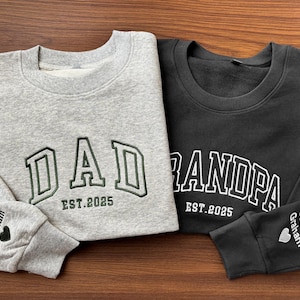 May include: Two gray and black sweatshirts with embroidered text. The gray sweatshirt says "DAD EST. 2025" and the black sweatshirt says "GRANDPA EST. 2025". Both sweatshirts have a small heart and the name "Cecilia Graham" embroidered on the sleeve.