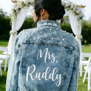 May include: Light blue denim jacket with pearl embellishments and the text "Mrs. Ruddy" in white script. The date "06.05.26" is embroidered on the collar. The jacket is a wedding-themed item.