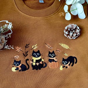 Peut inclure: Un sweat-shirt marron avec un motif brodé de quatre chats noirs avec des feuilles d'automne et des écharpes. Les chats sont entourés de feuilles brodées et d'une citrouille. Le sweat-shirt a un col rond.