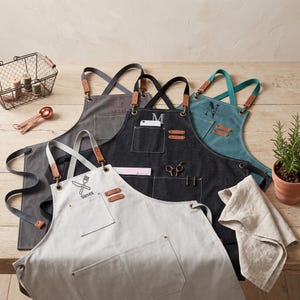 May include: Four canvas aprons in light gray, dark gray, black, and teal, each featuring cross-back straps with leather accents, multiple pockets, and personalized embroidery. The light gray apron displays a fork and knife icon with the name Steven.
