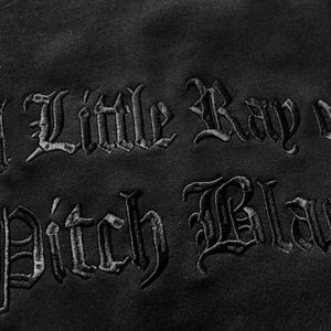 May include: Close-up of black fabric with the embroidered phrase "A Little Ray of Pitch Black" in a gothic font. The dark text is stitched onto the black material, creating a textured effect. The image highlights the contrast between the thread and the fabric.