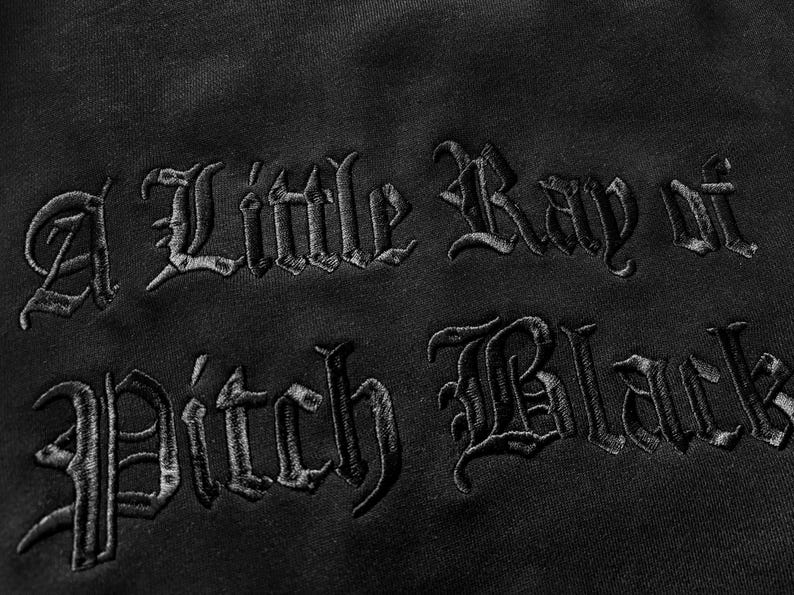 May include: Close-up of black fabric with the embroidered phrase "A Little Ray of Pitch Black" in a gothic font. The dark text is stitched onto the black material, creating a textured effect. The image highlights the contrast between the thread and the fabric.