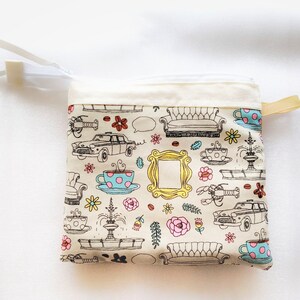 May include: A white zippered pouch with a Friends theme. The pouch is covered in a pattern of iconic Friends characters and objects, including the iconic orange couch, a coffee cup, a lobster, a fountain, and a yellow picture frame.