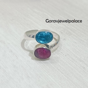 May include: A silver ring featuring two oval gemstones: a vibrant blue stone and a deep pink stone. The ring is set against a light-colored surface, with the text "Goravjewelpalace" visible.