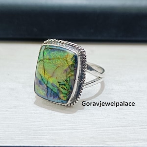 May include: A silver ring with a rectangular, iridescent gemstone. The stone displays shades of green, yellow, and blue. The ring has a braided border and a split shank. The text "Goravjewelpalace" is visible.