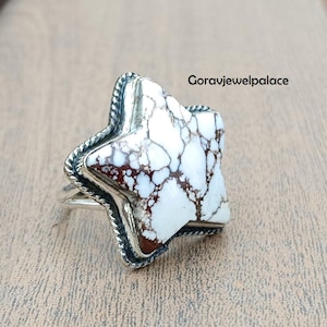 Könnte beinhalten: Ein sternförmiger Ring mit einem weißen und braunen Stein in einem silbernen Band. Der Stein hat ein einzigartiges Muster. Der Ring wird auf einer Holzoberfläche präsentiert. Ein auffälliges Schmuckstück.