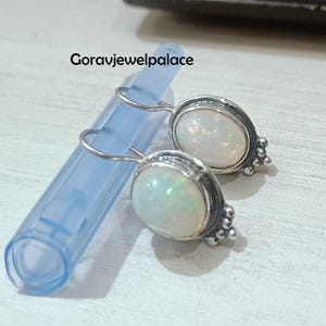 Op de afbeelding: Ovale opaal oorbellen met een zilverkleurige rand en een cluster van kleine kralen. De oorbellen worden getoond op een lichtblauwe plastic buis. De tekst "Goravjewelpalace" is zichtbaar.