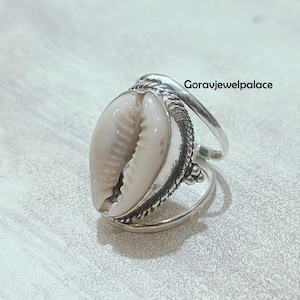 Könnte beinhalten: Ein Silberring mit einer natürlichen Kaurischnecke. Die Muschel hat eine cremeweiße Farbe und ist in einem detaillierten Silberband gefasst. Der Ring hat ein einzigartiges, organisches Design, ideal für Liebhaber von Schmuck im Strandstil. Der Text "Goravjewelpalace" ist sichtbar.