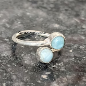 Könnte beinhalten: Ein silberner Ring mit zwei hellblauen, runden Edelsteinen. Der Ring hat ein schlichtes, offenes Design, wobei die Edelsteine an gegenüberliegenden Enden des Bandes angebracht sind. Der Ring wird auf einer dunklen, strukturierten Oberfläche präsentiert. Der Text "Goravjewelpalace" ist sichtbar.