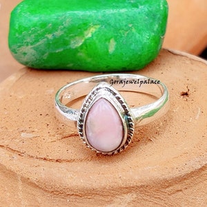 Peut inclure: Une bague en argent avec une pierre précieuse rose en forme de poire sertie dans une bélière en argent. La bague est posée sur une surface brune avec une pierre verte derrière elle. Le texte "GoraJewelpalace" est visible sur la bague.