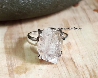 May include: A silver ring with a clear, faceted quartz stone. The stone is set in a claw setting and the ring is on a wooden surface.