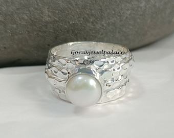 May include: A silver ring with a hammered texture and a white pearl set in the center. The ring is wide and has a textured band.