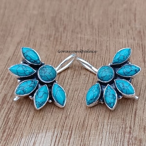 Puede incluir: Un par de pendientes de plata con piedras turquesa en forma de flor. Los pendientes están sobre una superficie de madera.