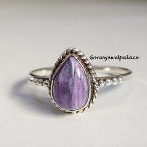 Puede incluir: Un anillo de plata con una piedra en forma de pera de color lavanda. La piedra está engastada en un borde trenzado en forma de cuerda. La banda del anillo tiene un diseño de cuentas en los lados. El texto "Goravjewelpalace" es visible.