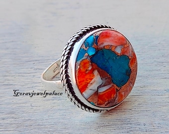 May include: A silver ring with a round, multi-colored stone. The stone is a mix of orange, white, and blue. The ring has a braided band.