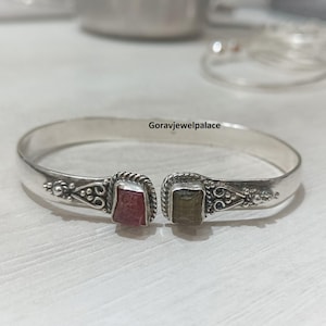 May include: A silver-toned cuff bracelet with decorative detailing and two square-cut gemstones, one red and one green. The bracelet is open-ended and rests on a light-colored surface. The text "Goravjewelpalace" is visible.