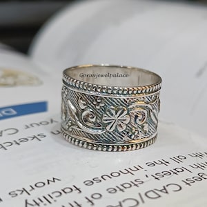 May include: A silver ring with an intricate floral design. The ring is wide and has a textured surface.
