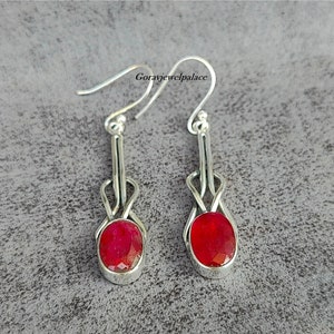 May include: A pair of silver dangle earrings with oval, faceted, red gemstones. The earrings have a unique design with a loop and a hook for the ear. The gemstones are set in a silver bezel.