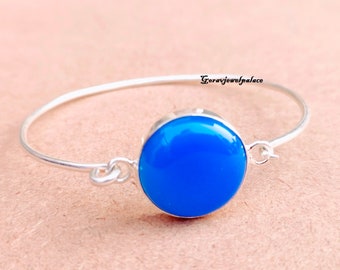May include: A silver bangle bracelet with a bright blue round cabochon gemstone. The bracelet is open and has a simple clasp.