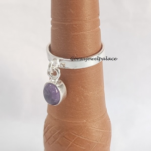 May include: A silver ring with a dangling charm featuring a purple gemstone. The ring is on a brown finger-sized display stand. The text "Goravjewelpalace" is visible on the ring.