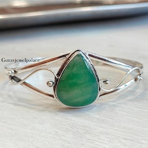 May include: A silver bangle bracelet featuring a teardrop-shaped green gemstone. The bracelet has a delicate, ornate design.